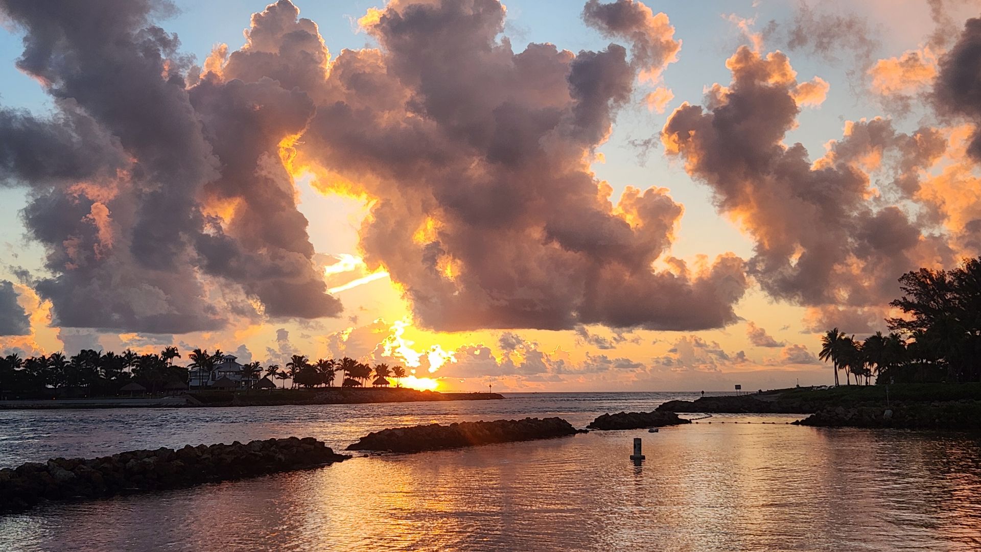 Sunrise over the Jupiter Inlet, with the jetty rocks in the foreground and palms silhouetted along the far shore.